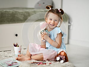 Happy kid girl in home dress sits on soft carpet at home hugging playing with dolls and drawing with colorful pencils