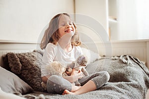Happy kid girl playing with teddy bears in her room, sitting on bed