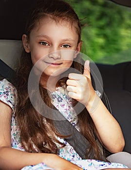 Happy kid girl sitting in the car with seat safety belt and showing thumb up by hand on summer green background