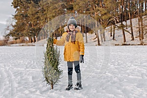 Happy kid holding in hands cut christmas tree on the background of snowy forest. Boy with xmas fir tree. Concept of winter