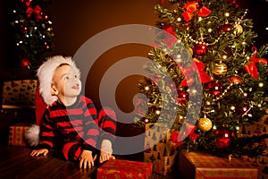 Happy Kid near Christmas Tree, Happy Baby Boy in Night Xmas Room, Child in Red Santa Hat, Present gift box