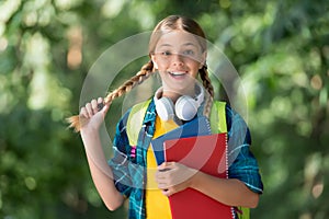 Happy kid with pigtails carry school books for foreign language learning wearing headphones natural landscape, English
