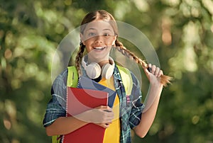 Happy kid with pigtails carry school books for foreign language learning wearing headphones natural landscape, English