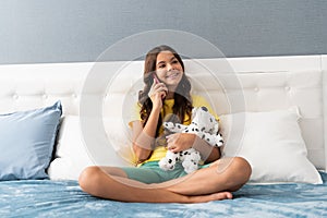 Happy kid talk on mobile phone sitting with toy puppy in bed, informal call