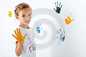 happy kid with yellow paint on hand near hand prints on white.