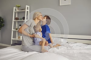 Happy kids congratulating mommy on Mother`s Day kissing her tenderly sitting on bed in bedroom