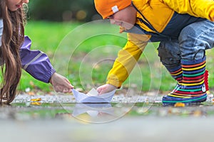 Happy kids girl and boy and colorful rubber boots playing with paper boat in puddle in autumn on nature