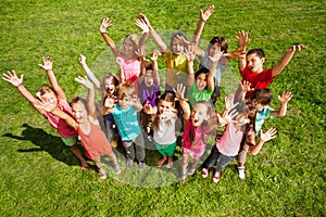 Grande gruppo di bambini felici, ragazzi e ragazze, alzate le mani per circa 10 anni, in piedi sull'erba verde vista dall'alto.