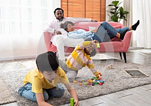 Happy kids playing toys and enjoying playtime activity while sitting on floor carpet in living room. Black couple of parents