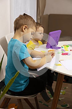 Happy kids with yellow and blue shirts doing arts and crafts together at their desk and watching cartoon on digital