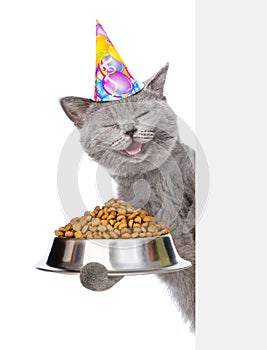 Happy kitten in birthday hat holding bowl of dry cat food and peeking from behind empty board. isolated on white background