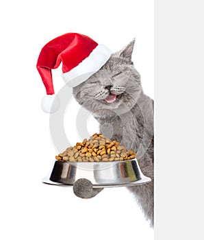 Happy kitten in red christmas hat holding bowl of dry cat food and peeking from behind empty board. isolated on white background