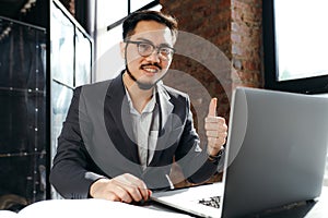 Happy korean businessman with glasses working at laptop and showing thumb up because of successful work