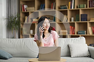 Happy korean woman using laptop and talking on cellphone at home, sitting on sofa, enjoying pleasant phone conversation
