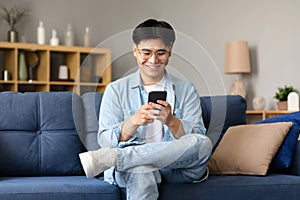 Happy Korean Young Man Using Smartphone Texting Sitting At Home