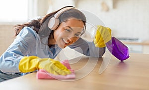 Happy lady dusting table cleaning desktop with rag caring for wooden furniture at home and listening music in headphones