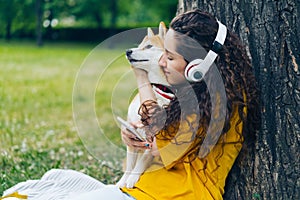 Happy lady in headphones using smartphone and hugging pet dog in park