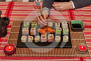 Happy lady holds a sushi roll on the chopsticks, looks at delicious Japanese food and smiles, over a table sit on a sofa