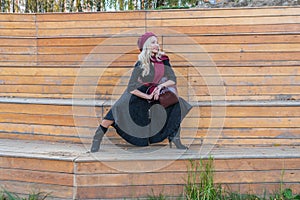 Happy lady sits on a bench of a summer theater, wooden in a burgundy coat and biret, looking at the camera, in the fall