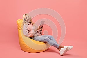 Happy lady using digital tablet while sitting in beanbag chair over pink background with free space