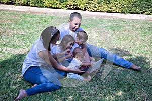 Happy, large family of five sitting on the grass in a park looking at a tablet. With three children, one of them a transsexual