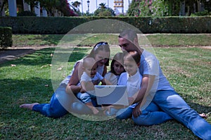 Happy, large family of five sitting on the grass in a park looking at a tablet. With three children, one of them a transsexual
