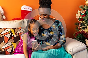 Happy Latin Afro-descendant Colombian mother and daughter sitting on the sofa next to the Christmas tree checking the cell phone