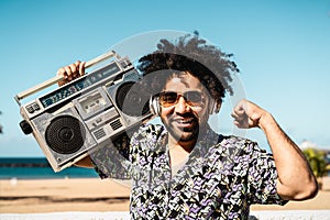 Happy Latin man having fun listening to music with wireless headphones and vintage boombox on the beach during summer