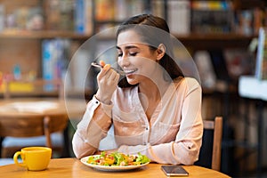 Happy latin woman eating lunch in cafe, enjoying delicious salad with closed eyes and drinking hot beverage.