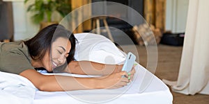 Happy laughing Asian woman lies in bed with mobile phone. She received a declaration of love from her boyfriend. Format