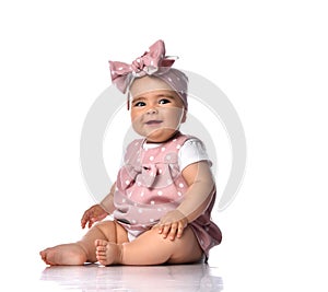 Happy laughing a bit sly infant baby girl in polka dot dress and headband with bow sits on the floor holding her knee