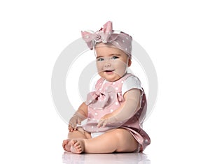 Happy laughing a bit sly infant baby girl in polka dot dress and headband with bow sits on the floor looking at camera