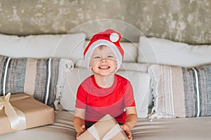Happy laughing Caucasian boy in red Santa Claus hat with his presents on Christmas morning. Child excited and smiling