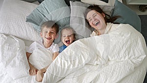 Happy laughing family with two kids hiding under blanket in bed and throwing it off. Concept of family happiness.