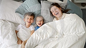 Happy laughing family with two kids hiding under blanket in bed and throwing it off. Concept of family happiness, relaxing at home