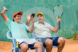 Happy laughing indian senior friends at tennis court by watching mobile phone - concept of taking break, social media