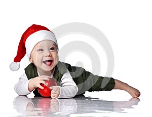 Happy laughing infant boy toddler in red Santa hat plays with Christmas New Year fir tree ball on floor looking aside