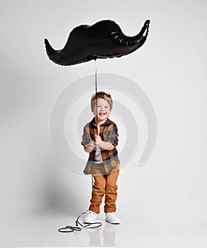 Happy laughing kid boy in jeans and shirt having fun playing with big moustache air balloon, jumping, trying to fly