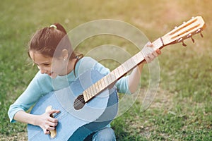 Happy laughing little girl in blue blouse playing songs on custom made denim acoustic guitar sitting on green grass outdoors