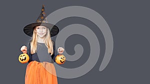 A happy laughing little girl in a carnival witch costume holds a Halloween pumpkin baskets for treats on a dark
