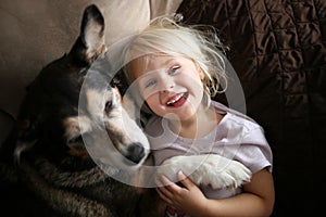 Happy, Laughing Little Girl Child Hugging Pet Dog on Couch