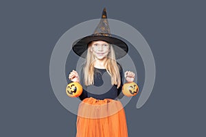 A happy laughing little girl in a witch costume and makeup holds pumpkin baskets for Halloween treats
