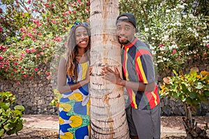 Happy laughing playful african millennial spouses posing hugging a palm tree, smiling and looking at camera, siblings, brother and