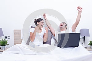 Happy laughing relaxed young couple using laptop in bed at home. they are celebrating. Intermet and work at home concept