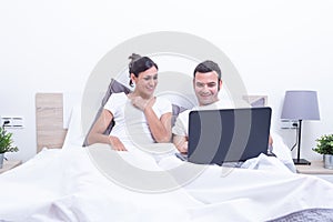 Happy laughing relaxed young couple using laptop in bed at home. Intermet and work at home concept