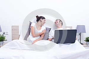 Happy laughing relaxed young couple using laptop in bed at home. Intermet and work at home concept