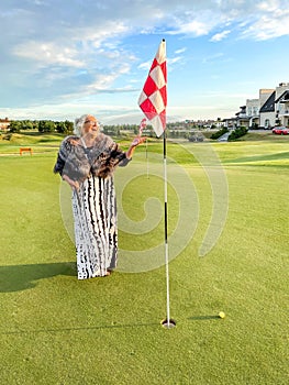 Happy laughing senior woman plays golf on golf course in evening attire. Ball is near hole with flag