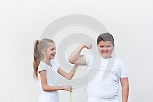 Happy laughing small caucasian slim girl measuring thick smiling kid muscle with green tape