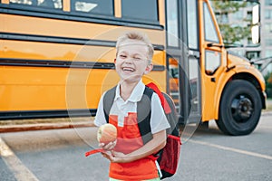 Happy laughing smiling Caucasian boy student by yellow bus on first September day. Hard of hearing child kid eating apple fruit at