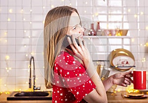 Happy laughing and smiling young beautiful woman talking by mobile phone in kitchen in red pygamas holding tea cup. New year and